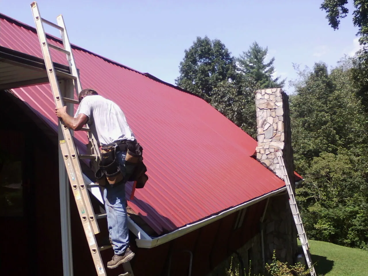 Roofer on a ladder beside a red metal roof and stone chimney — Soffit & Fascia Repair by Roofing Brothers in Clatonia